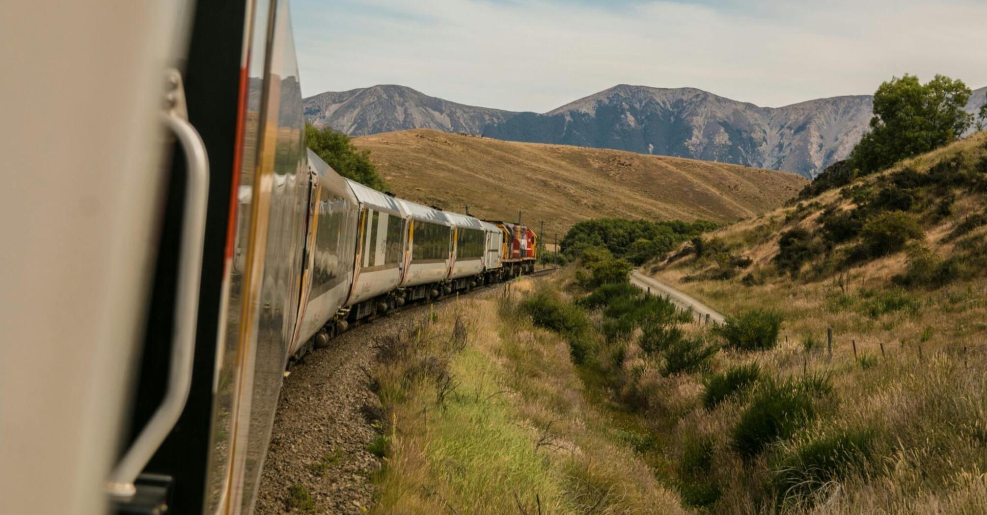 A passenger train traveling through scenic hills with mountains in the background under a clear sky