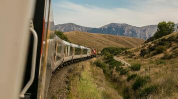 A passenger train traveling through scenic hills with mountains in the background under a clear sky