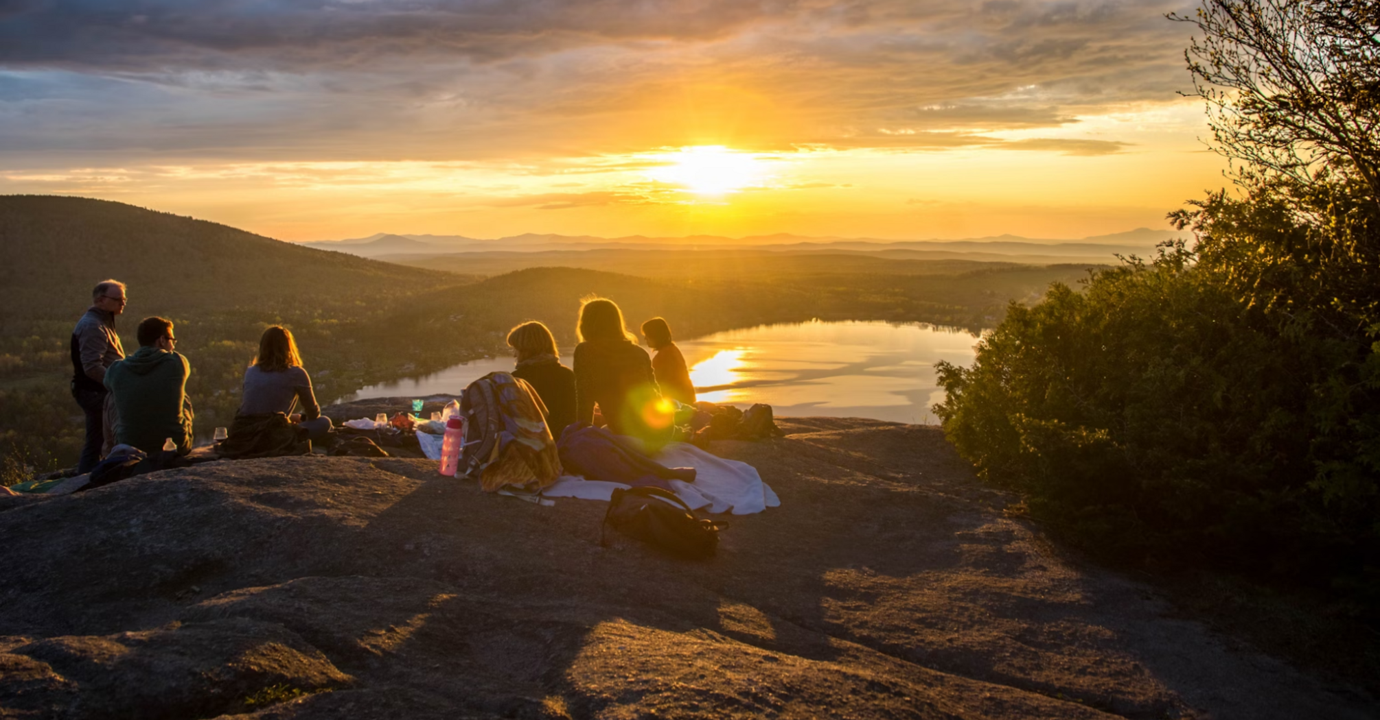 A group of people sitting on a mountain top during sunset, with a lake