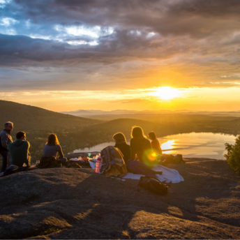 A group of people sitting on a mountain top during sunset, with a lake