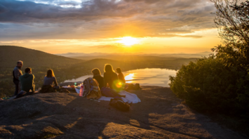 A group of people sitting on a mountain top during sunset, with a lake