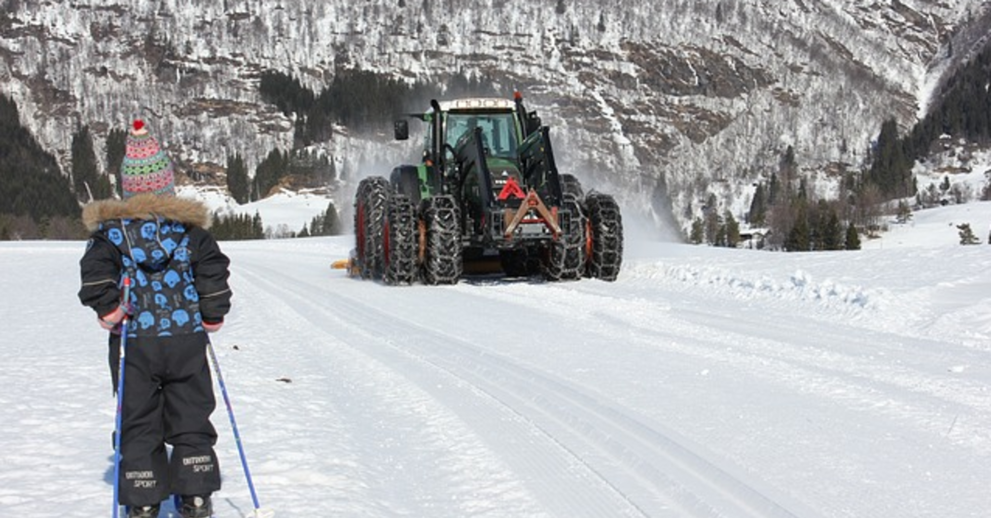 A child skiing towards a large snow tractor with six wheels, wrapped in chains for snow navigation