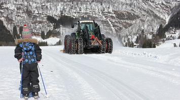 A child skiing towards a large snow tractor with six wheels, wrapped in chains for snow navigation