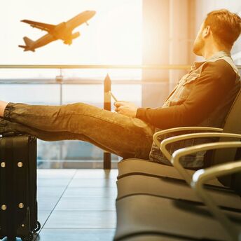 A traveler relaxing at an airport terminal with legs resting on a suitcase, watching a plane take off through the window