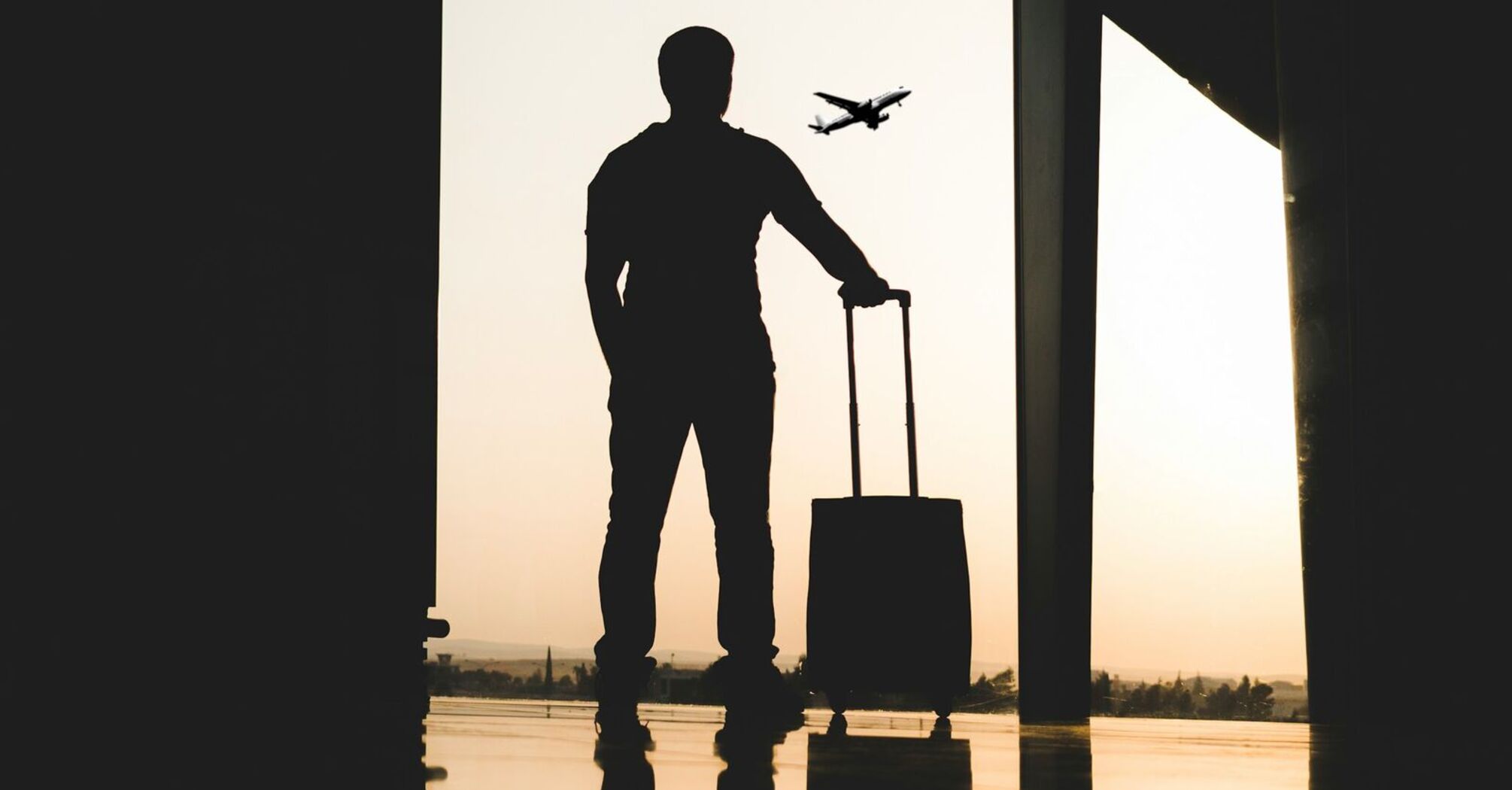 A silhouette of a traveler with a suitcase at an airport, watching a plane take off against the sunset