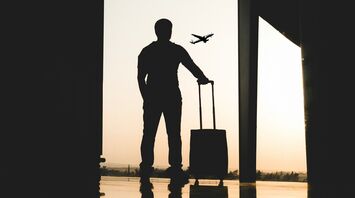 A silhouette of a traveler with a suitcase at an airport, watching a plane take off against the sunset