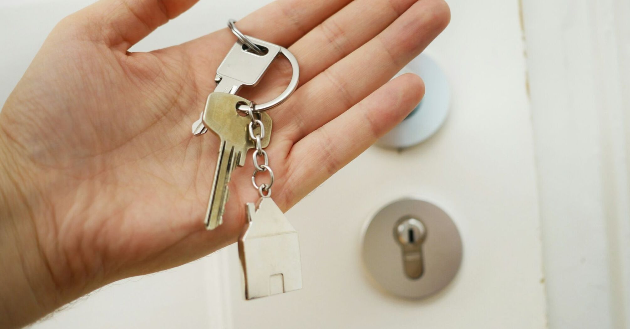 A hand holding a set of house keys with a keychain in the shape of a house, in front of a door lock