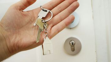 A hand holding a set of house keys with a keychain in the shape of a house, in front of a door lock