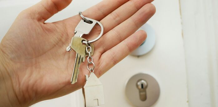 A hand holding a set of house keys with a keychain in the shape of a house, in front of a door lock