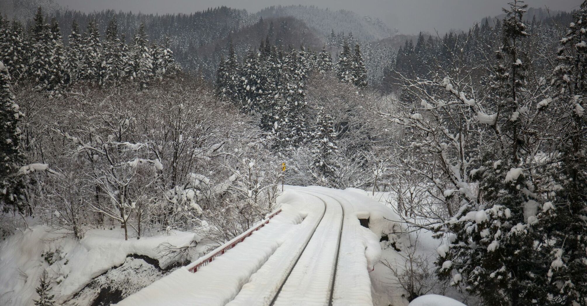 A snow-covered railway track passing through a forested area during winter, with trees and hills blanketed in snow