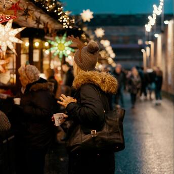 Woman holding a cup at a Christmas market, surrounded by festive lights and decorated stalls in the evening