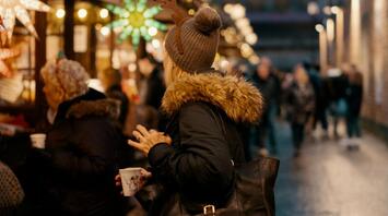 Woman holding a cup at a Christmas market, surrounded by festive lights and decorated stalls in the evening
