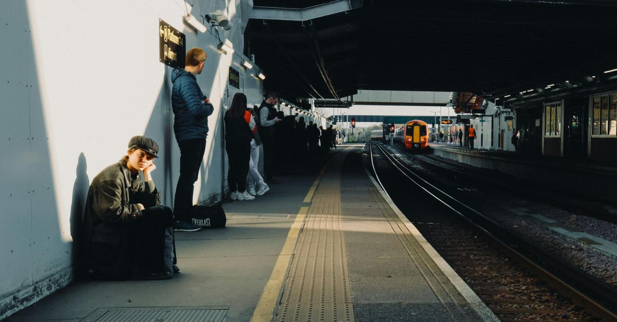 Passengers waiting at a train station platform in sunlight as a train approaches