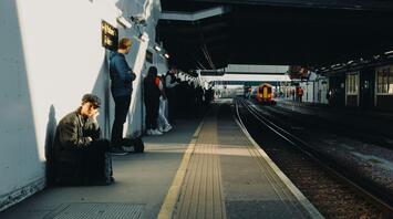 Passengers waiting at a train station platform in sunlight as a train approaches
