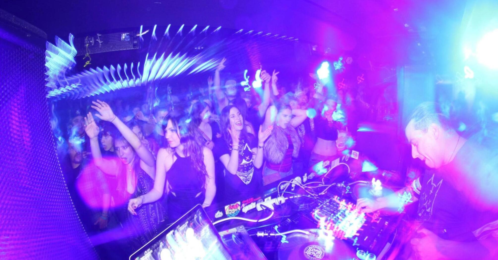 Crowded nightclub with a DJ performing under bright purple and blue lights