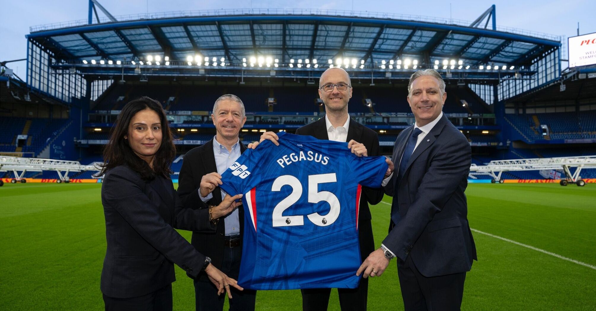 Four people hold a Chelsea football shirt with “Pegasus 25” on the pitch of an empty stadium