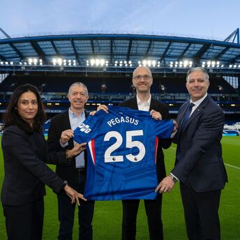 Four people hold a Chelsea football shirt with “Pegasus 25” on the pitch of an empty stadium