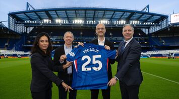 Four people hold a Chelsea football shirt with “Pegasus 25” on the pitch of an empty stadium