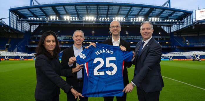 Four people hold a Chelsea football shirt with “Pegasus 25” on the pitch of an empty stadium