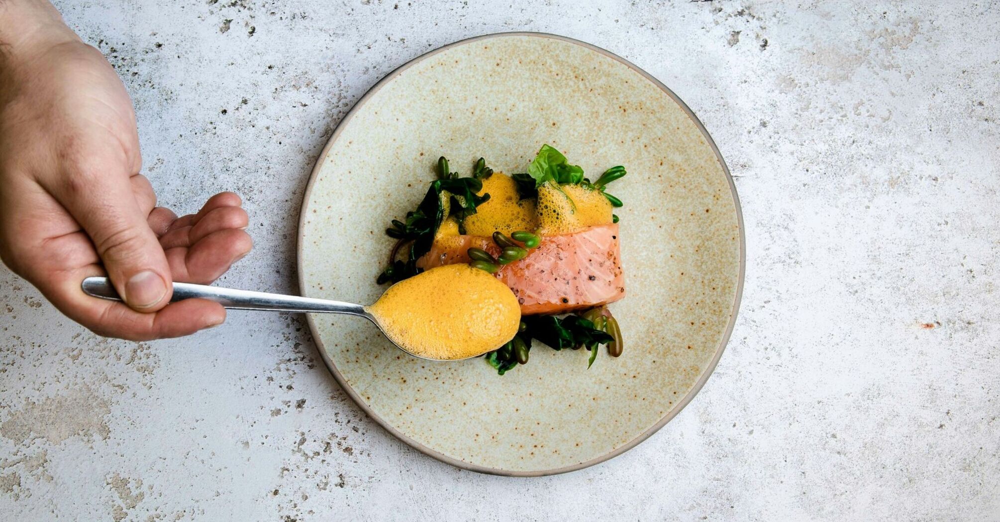 A plated gourmet dish with salmon, greens, and foam being carefully arranged by a chef