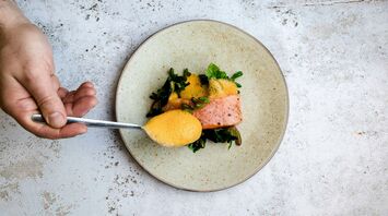 A plated gourmet dish with salmon, greens, and foam being carefully arranged by a chef