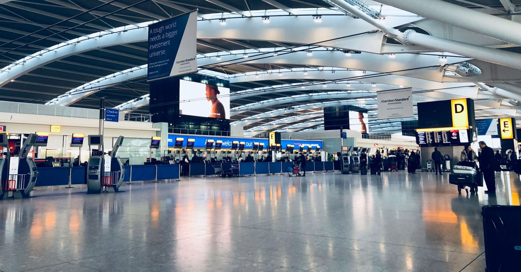 A modern, spacious terminal at Heathrow Airport with check-in kiosks, passengers, and illuminated signage