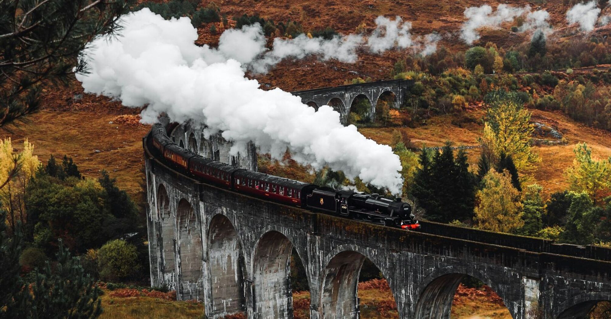 Steam train crossing the iconic Glenfinnan Viaduct in the Scottish Highlands, surrounded by autumn foliage