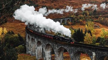Steam train crossing the iconic Glenfinnan Viaduct in the Scottish Highlands, surrounded by autumn foliage
