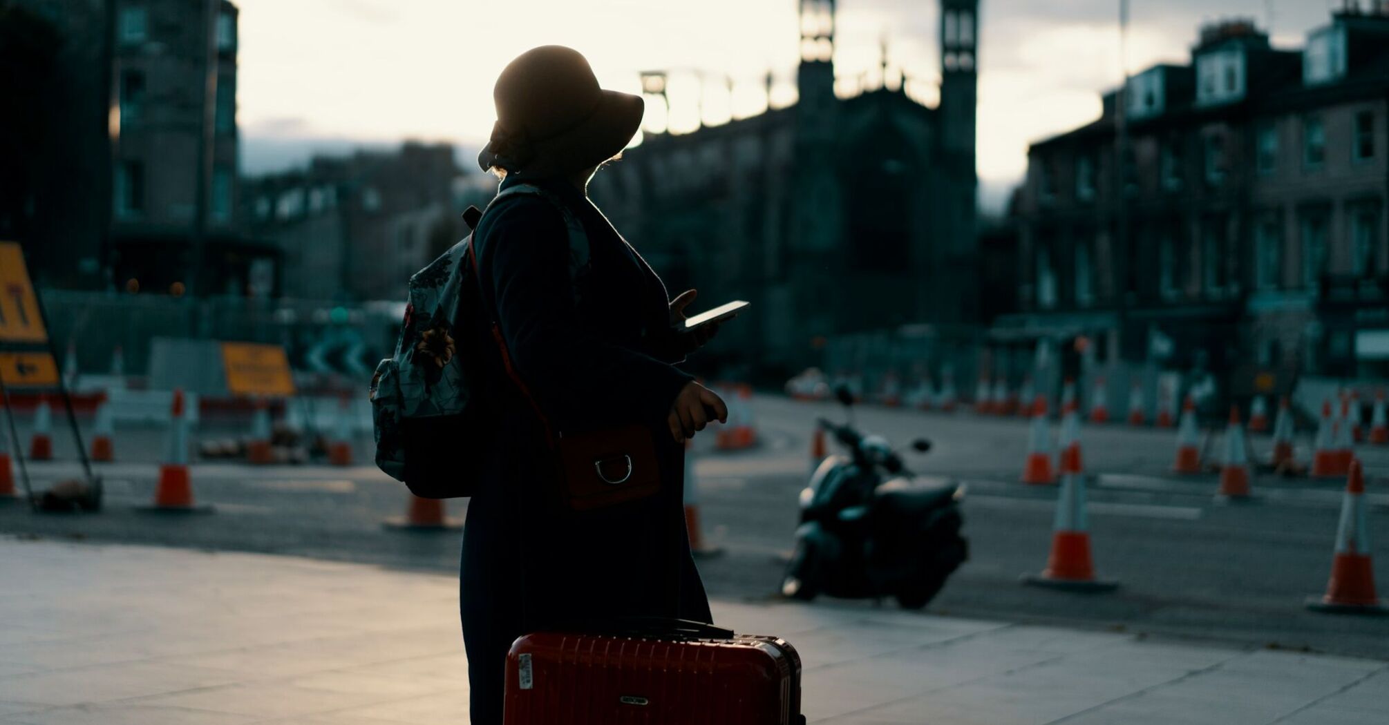 A silhouette of a traveler with a suitcase and smartphone standing near a construction site at dusk