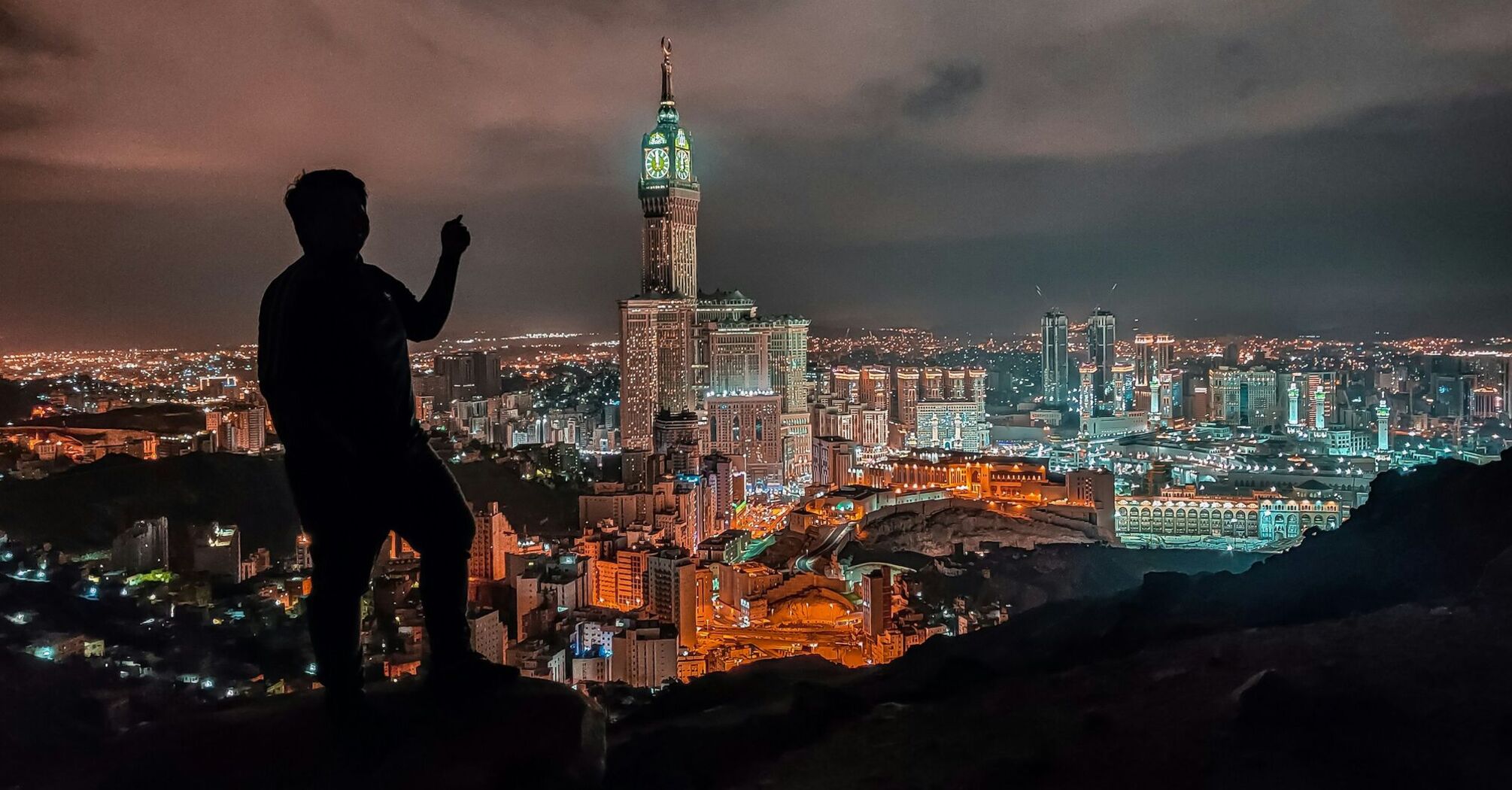 Silhouette of a person overlooking the illuminated skyline of Makkah at night, featuring the Abraj Al Bait Towers and the Grand Mosque