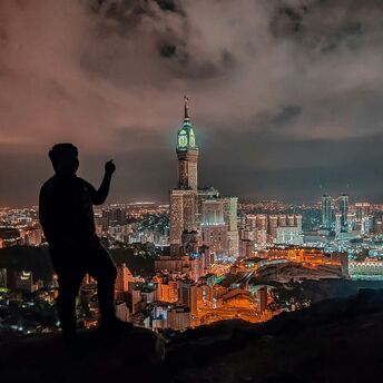 Silhouette of a person overlooking the illuminated skyline of Makkah at night, featuring the Abraj Al Bait Towers and the Grand Mosque