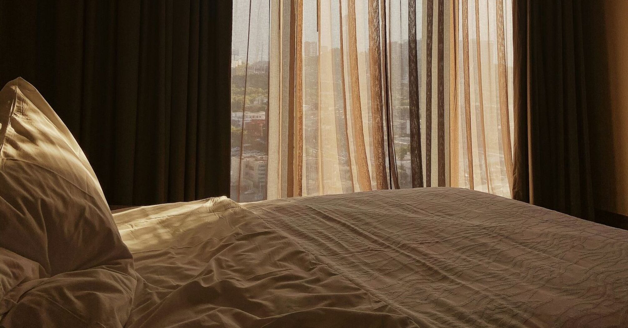 A cozy hotel room with sunlight filtering through sheer curtains, overlooking a cityscape