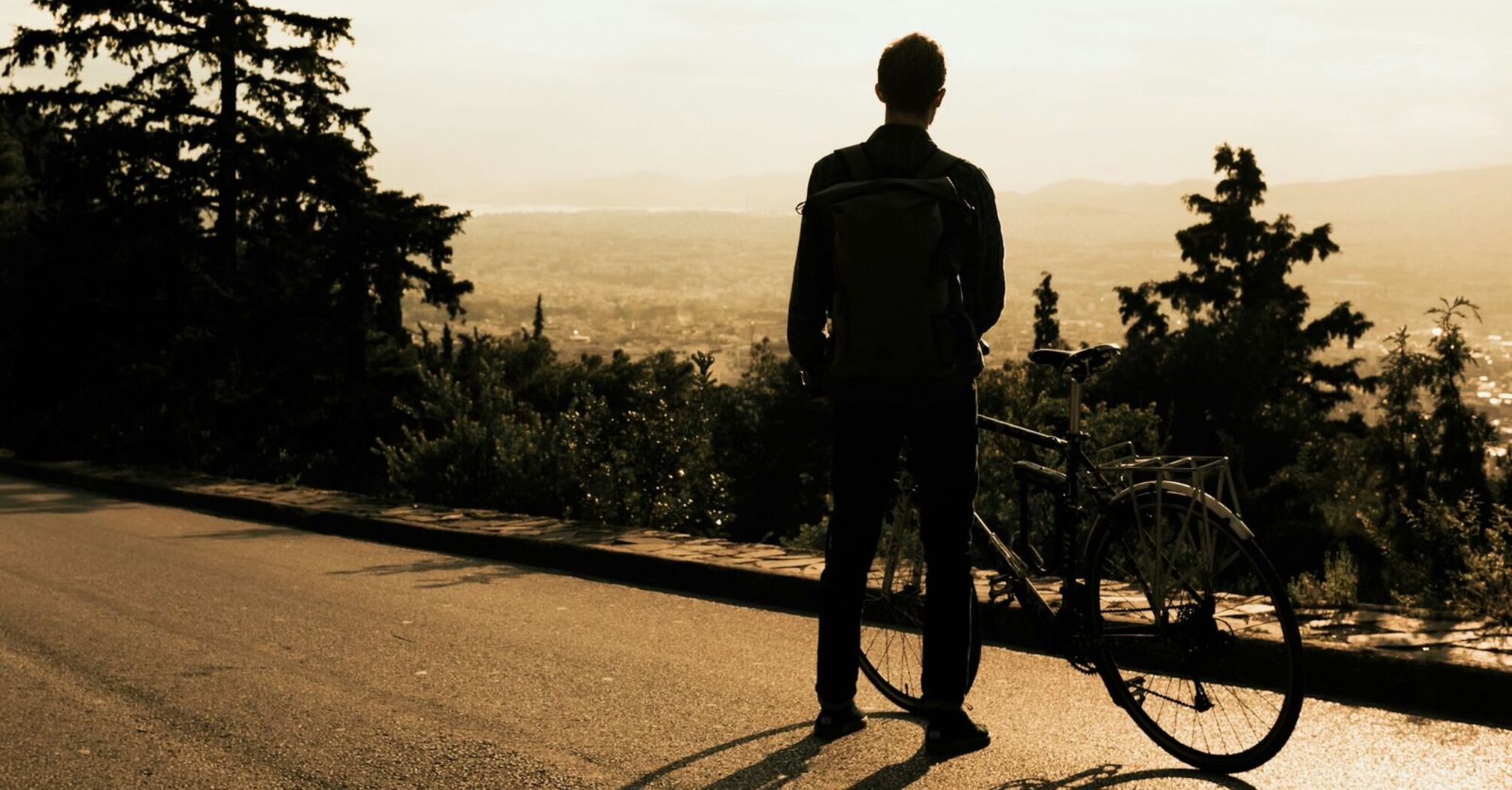 A cyclist standing with their bicycle on a scenic road, gazing at the sunset over a valley