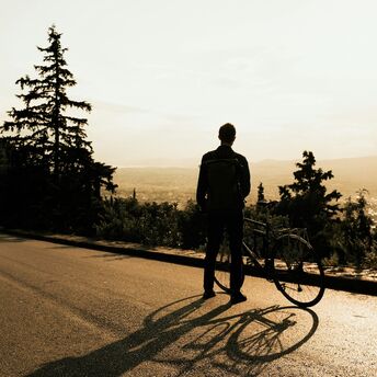 A cyclist standing with their bicycle on a scenic road, gazing at the sunset over a valley