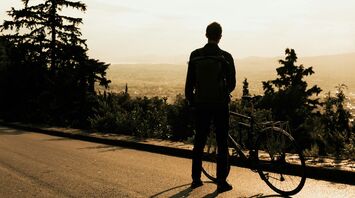 A cyclist standing with their bicycle on a scenic road, gazing at the sunset over a valley