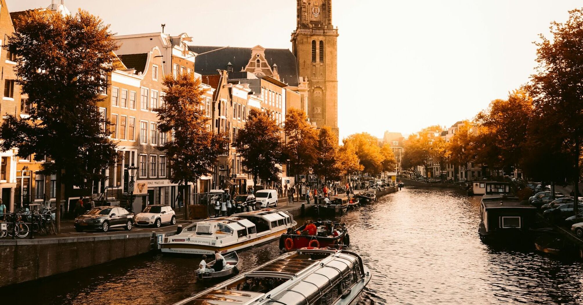 A scenic canal in Amsterdam with a glass-roofed tour boat and the historic Westerkerk tower in the background during golden hour