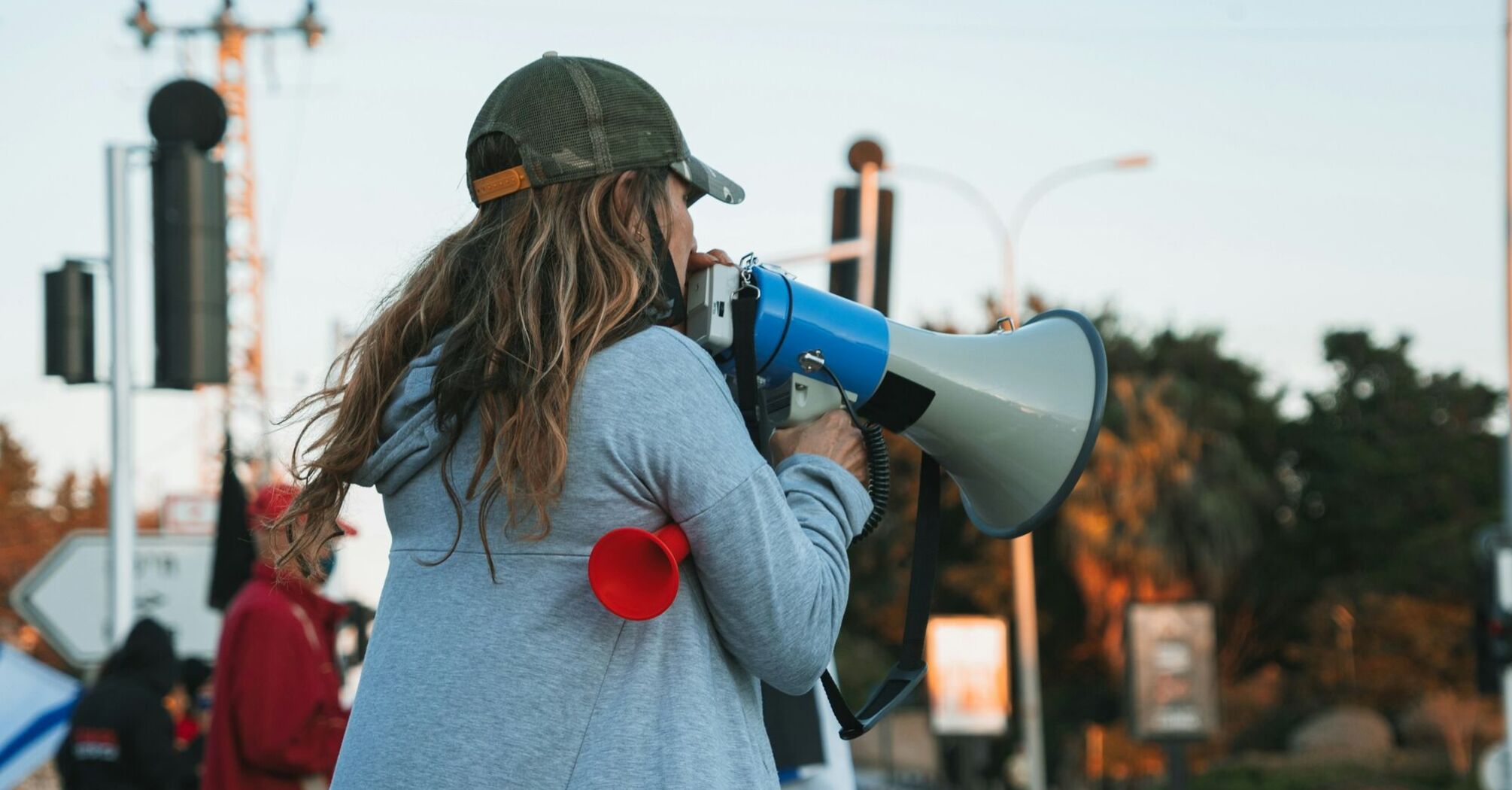 A woman in a gray hoodie and cap speaks through a megaphone at a protest, with other demonstrators in the background