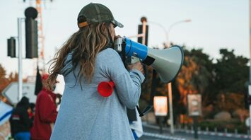 A woman in a gray hoodie and cap speaks through a megaphone at a protest, with other demonstrators in the background