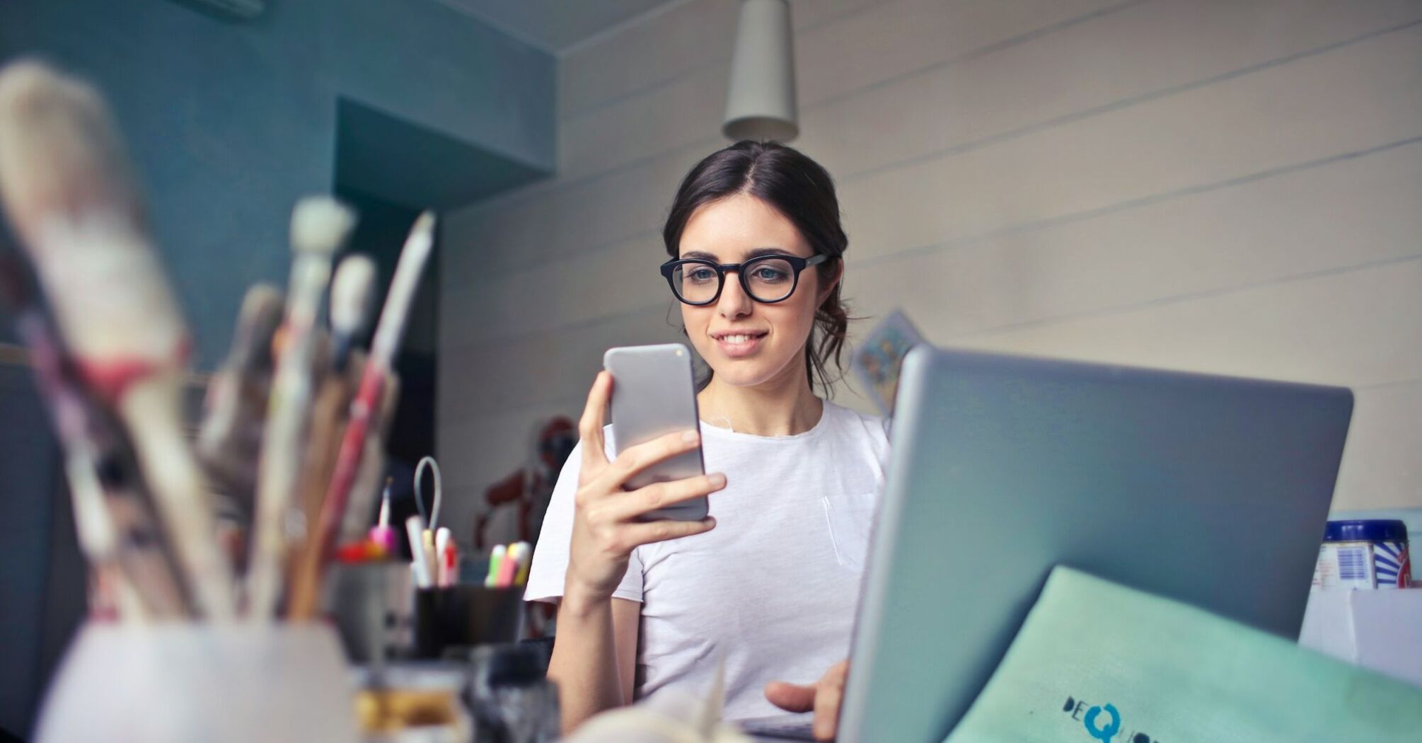 A young woman wearing glasses, using a smartphone while working on a laptop at a creative workspace with art supplies