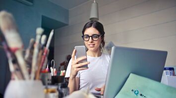 A young woman wearing glasses, using a smartphone while working on a laptop at a creative workspace with art supplies