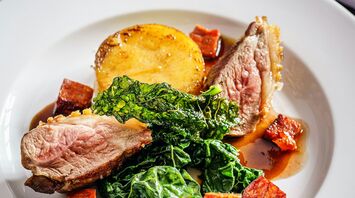 A gourmet dish featuring seared lamb, crispy kale, roasted vegetables, and a rich sauce on a white plate