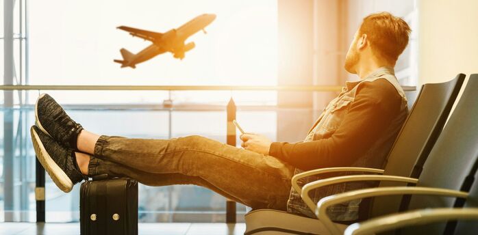 A traveler sitting in an airport lounge with legs resting on a suitcase, watching a plane take off in the background