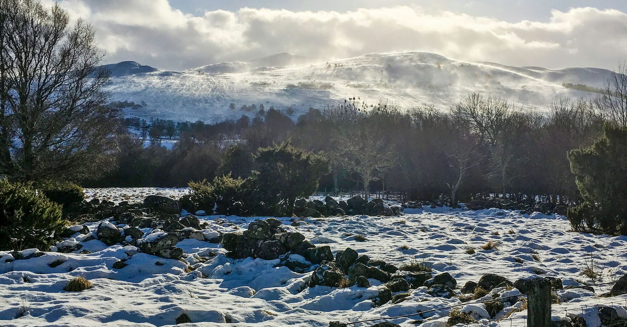 A snow-covered landscape in Scotland with hills in the background and scattered rocks in the foreground under a partly cloudy sky