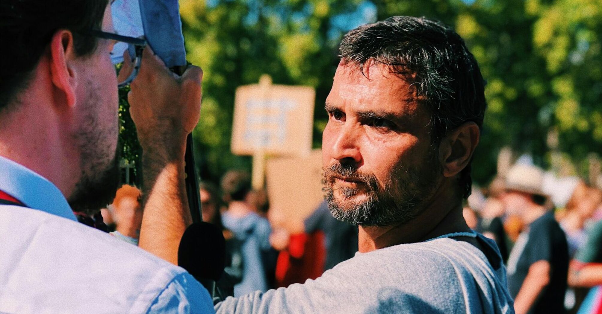 A determined man with a beard at a protest, engaging in conversation with another participant, with a crowd holding signs in the background