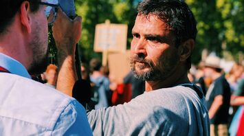 A determined man with a beard at a protest, engaging in conversation with another participant, with a crowd holding signs in the background