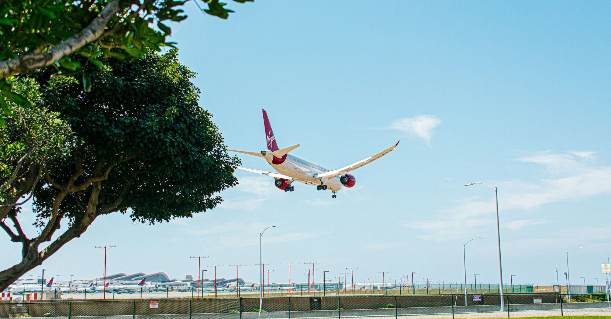 A Virgin Atlantic aircraft descends for landing at an airport on a clear day, with a green park in the foreground and multiple planes parked in the background