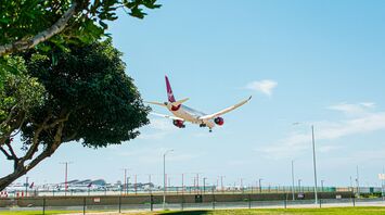 A Virgin Atlantic aircraft descends for landing at an airport on a clear day, with a green park in the foreground and multiple planes parked in the background