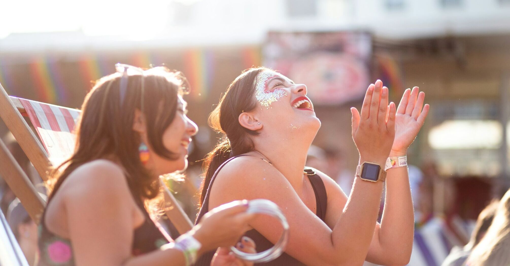 Two women enjoying a festival, laughing and clapping in the sunlight, with rainbow decorations in the background