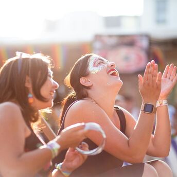 Two women enjoying a festival, laughing and clapping in the sunlight, with rainbow decorations in the background