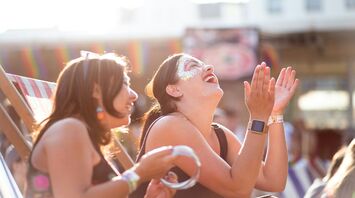 Two women enjoying a festival, laughing and clapping in the sunlight, with rainbow decorations in the background